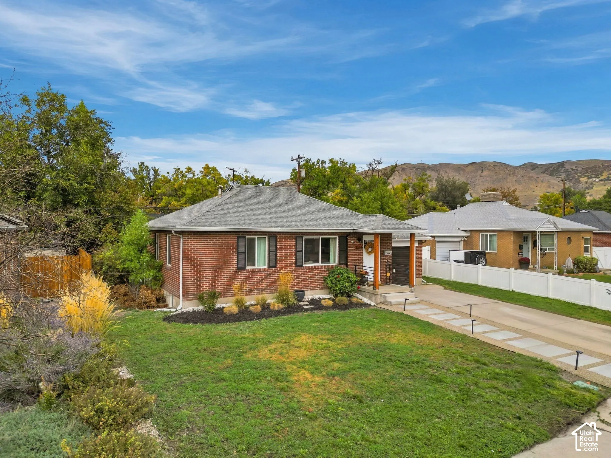 Brick exterior with dark shutters, landscaped front yard, gray shingle roof, paved driveway, and mountain backdrop