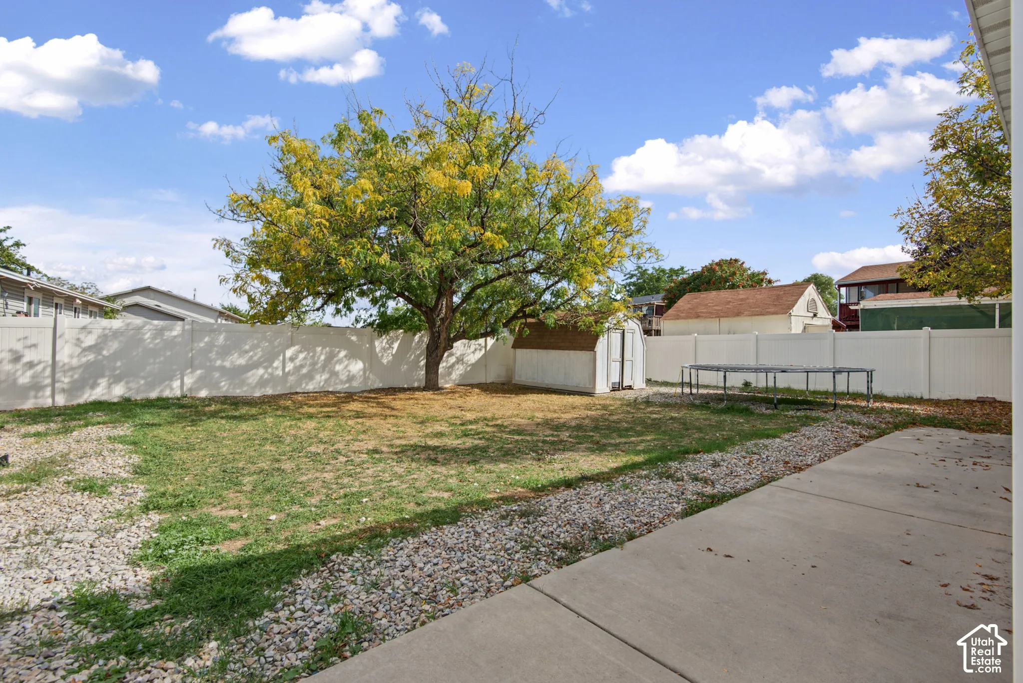 Expansive backyard featuring a mature deciduous tree, a storage shed with a shingled roof, a paved patio, and white privacy fencing