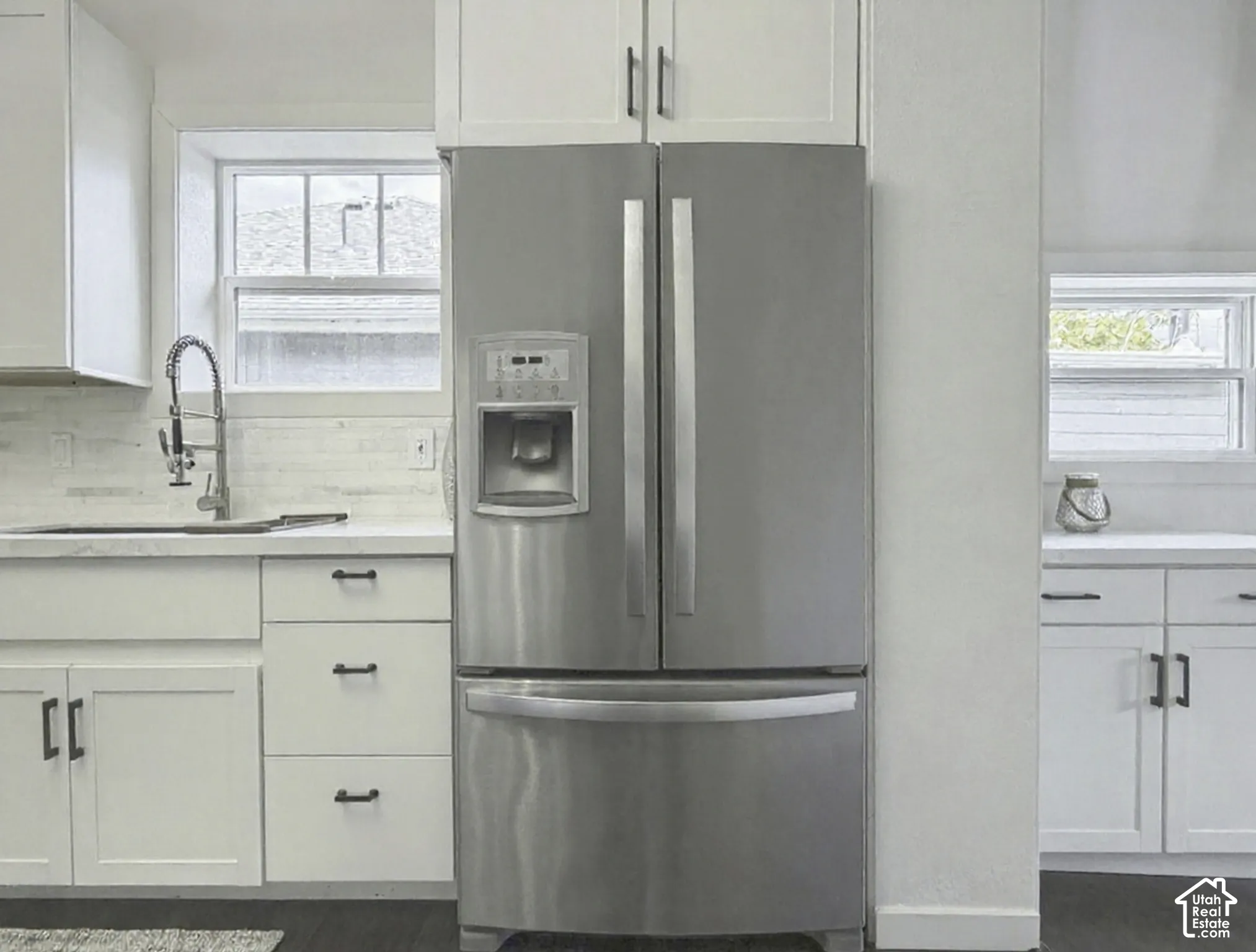 Stainless steel French door refrigerator, white shaker-style cabinetry with matte black hardware, dual windows, white countertops, and subway tile backsplash