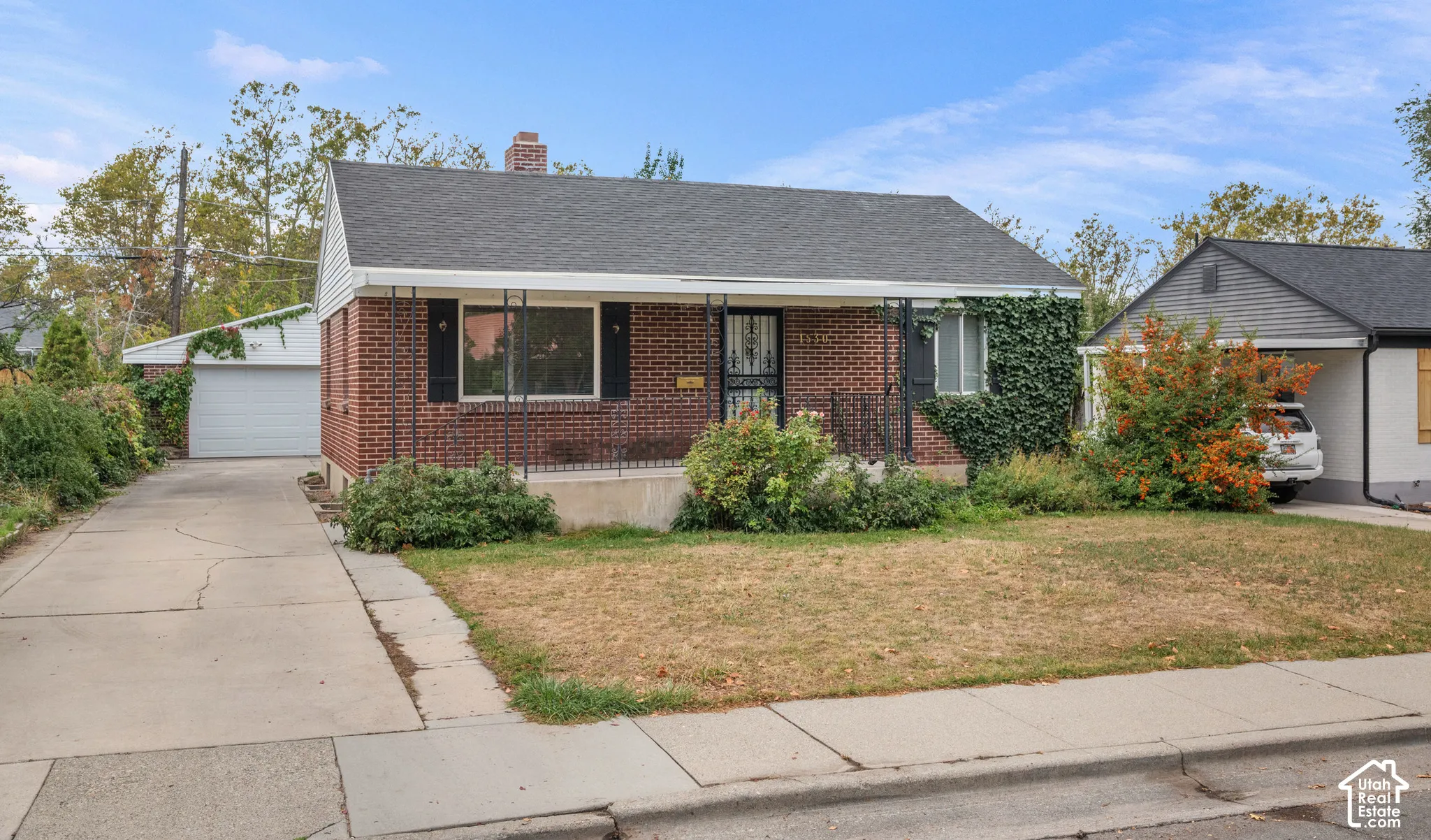 Brick exterior with a covered front porch, an attached garage, a paved driveway, and mature landscaping