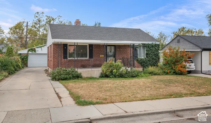 Brick exterior with a covered front porch, an attached garage, a paved driveway, and mature landscaping