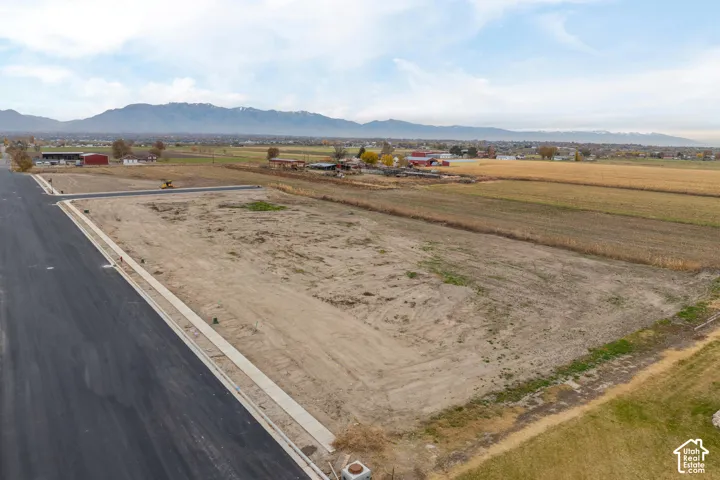 Paved road frontage, concrete sidewalks, bare land, mountain range backdrop, and agricultural fields