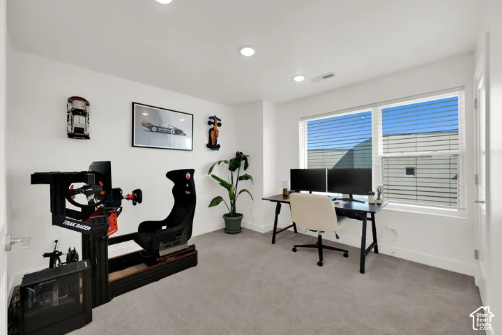 Recessed lighting illuminates a spacious room featuring a large window with blinds, light gray carpeting, and white walls