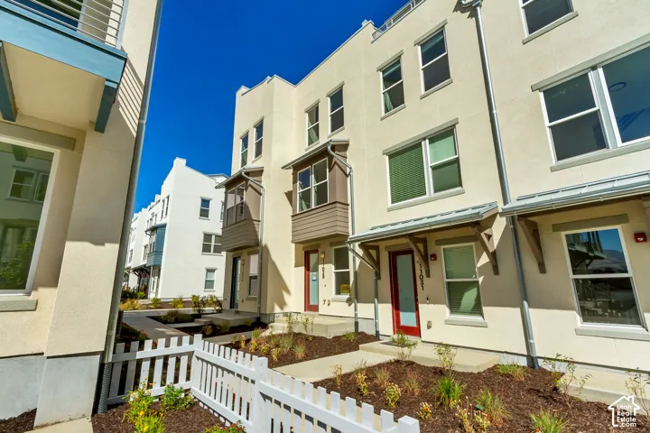 Contemporary townhome featuring a clean cream and white exterior with multi-pane windows, a recessed entry with red accent door, and a welcoming front facade with white picket fencing and newly landscaped planting beds