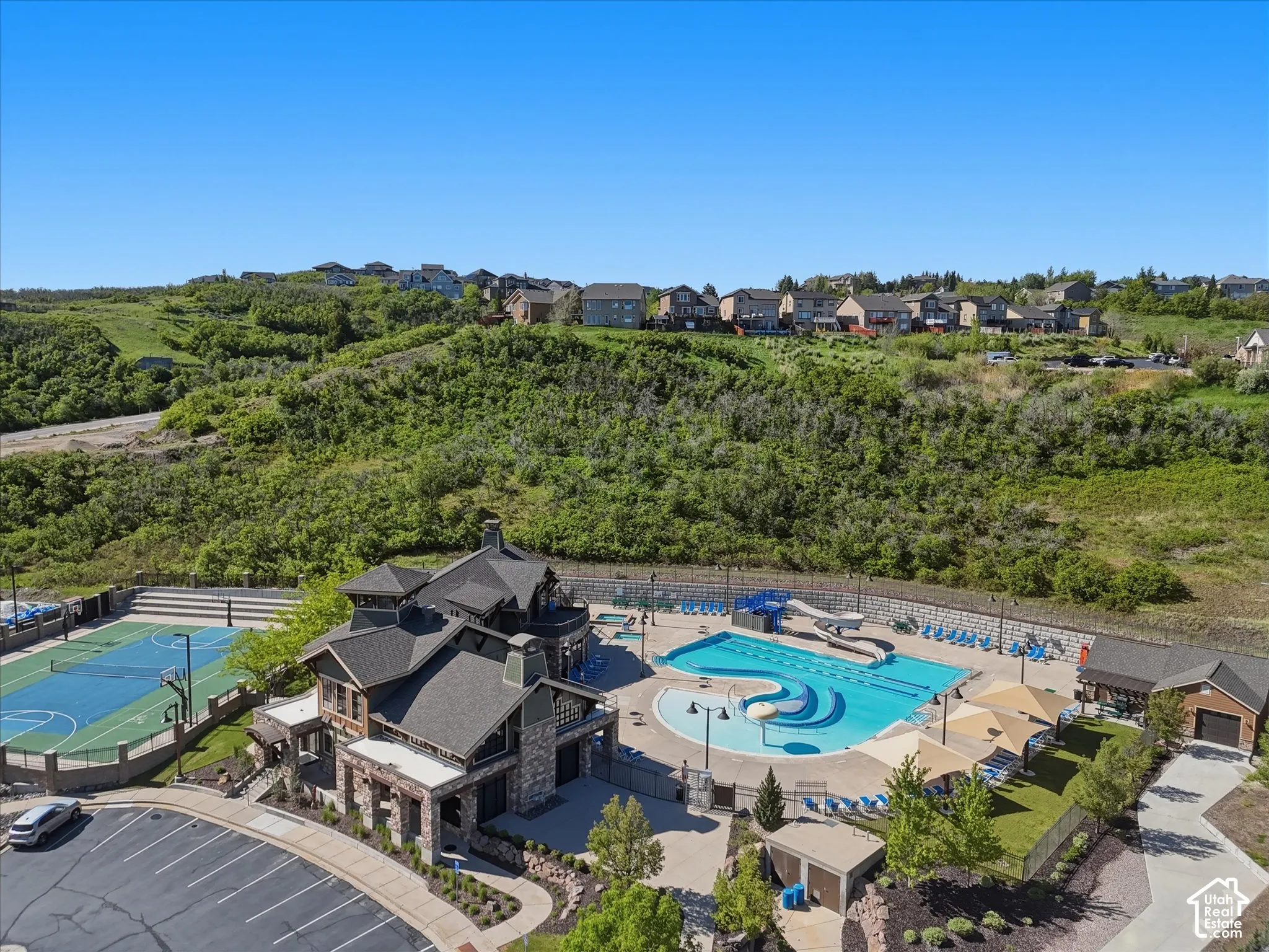 Community recreation center featuring a multi-sport court, expansive swimming pool with water slides, structured parking, and surrounding natural landscaping