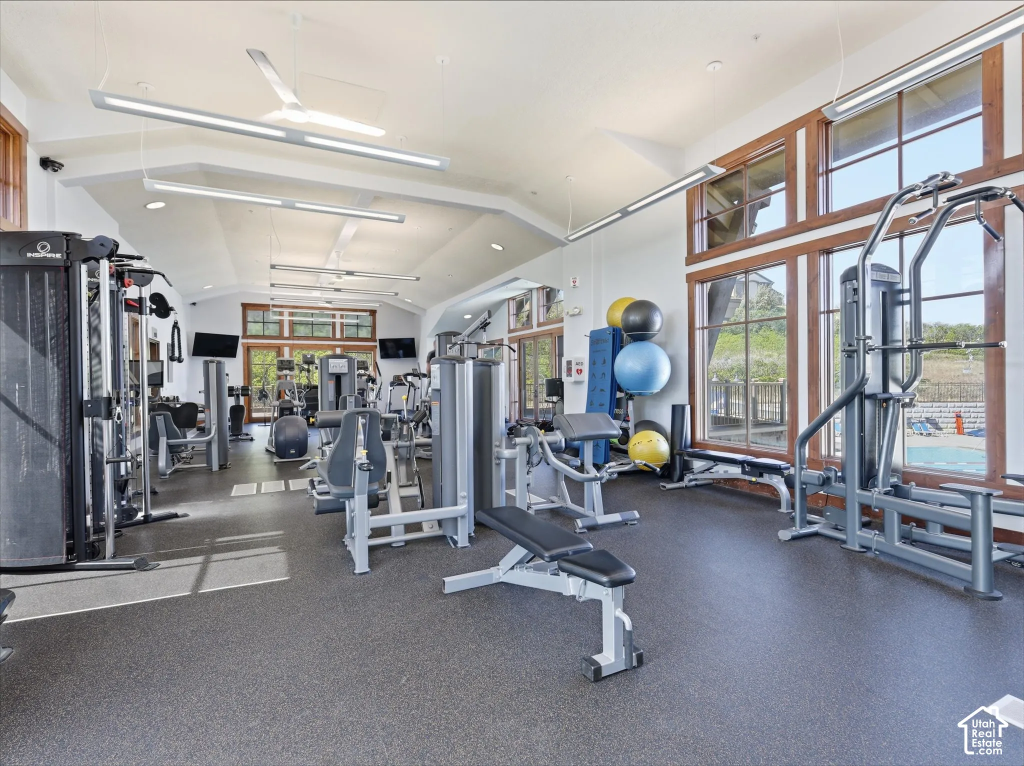 Vaulted ceiling with exposed wood beams, expansive window array with wood trim, durable rubber flooring, modern linear lighting fixtures, and ceiling fans