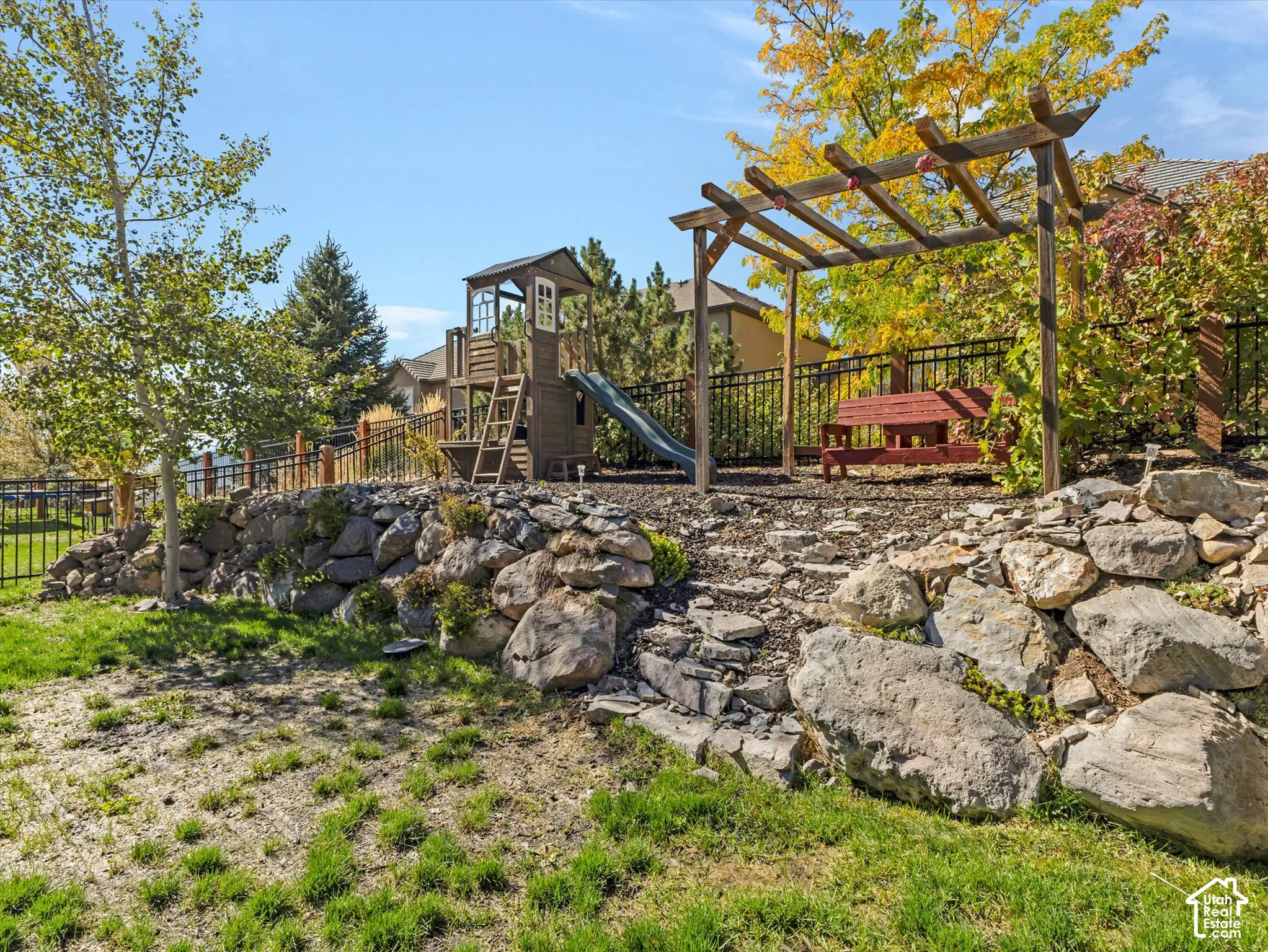 Terraced backyard featuring a rock retaining wall, mature trees, a wooden pergola, and a playset with a slide