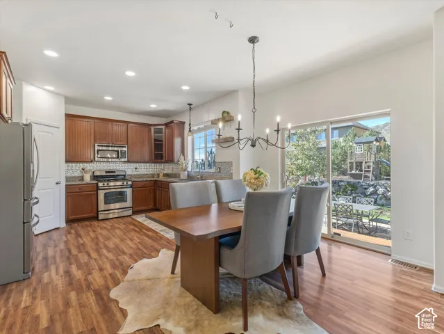 Open-concept kitchen and dining area featuring wood cabinetry, stainless steel appliances, granite countertops, wood-finish flooring throughout, and expansive glass sliding doors opening to outdoor space with mature landscaping
