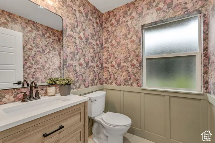 Floral wallpaper with wainscoting, wood-finish vanity with white countertop, integrated sink, and bronze-finish faucet