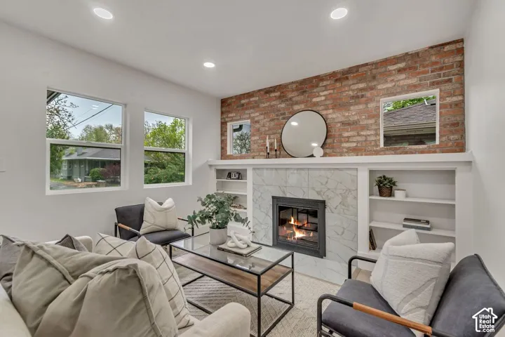 Sophisticated living room featuring exposed brick accent wall, gas fireplace with marble surround and built-in shelving, recessed ceiling lights, large multi-pane windows with natural light, and contemporary furnishings arrangement anchored by a glass-top