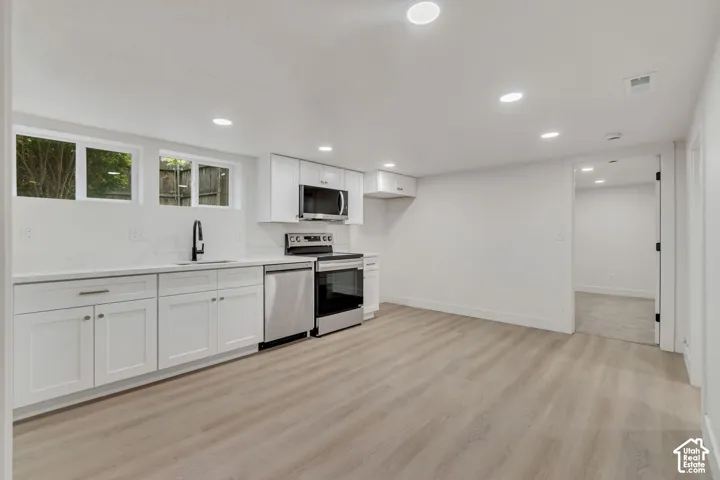 Modern kitchen and living space featuring white cabinetry with hardware accents, stainless steel appliances including a gas range and built-in microwave, wood-finish flooring throughout, recessed ceiling lighting, and large windows providing natural light