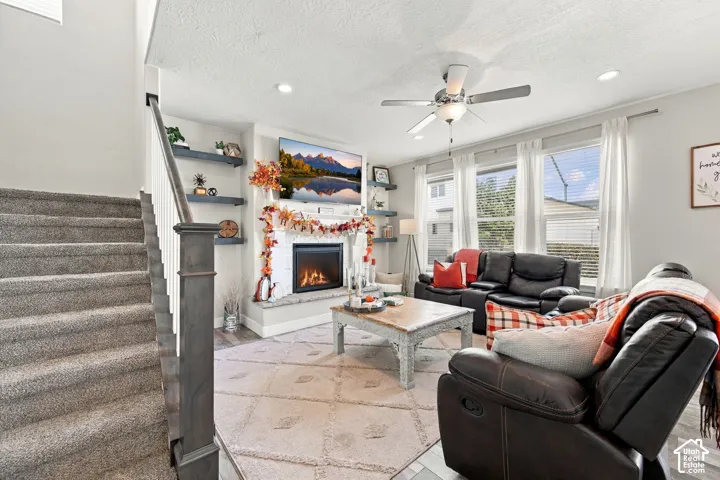 Living area featuring a fireplace with a stone hearth, built-in shelving, wood-finish flooring, a ceiling fan, and multiple windows with blinds