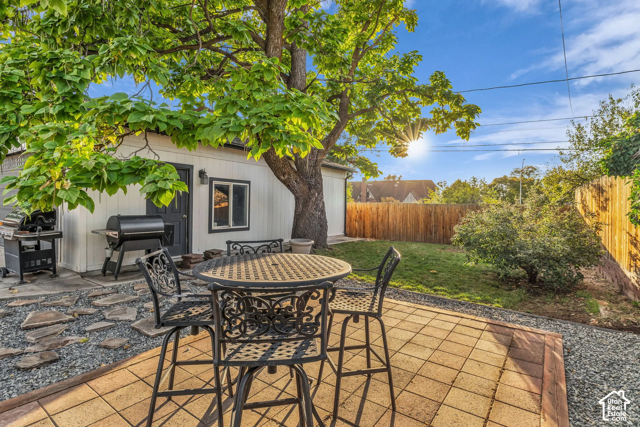 Outdoor entertaining space featuring a tile-paved patio with wrought iron dining set, mature shade tree, built-in grill, and wooden privacy fencing