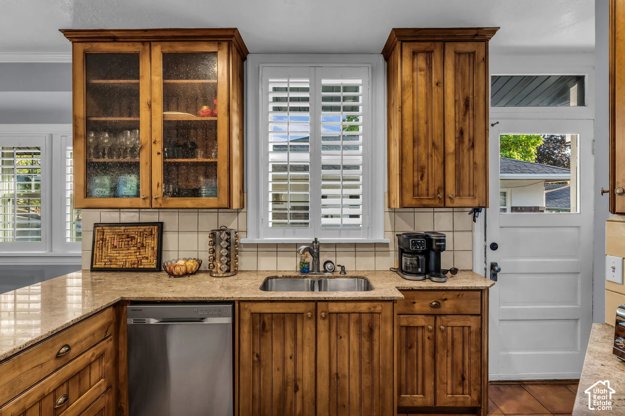 Wood-finish cabinetry with glass-front uppers, stainless steel dishwasher, integrated sink, and plantation shutters