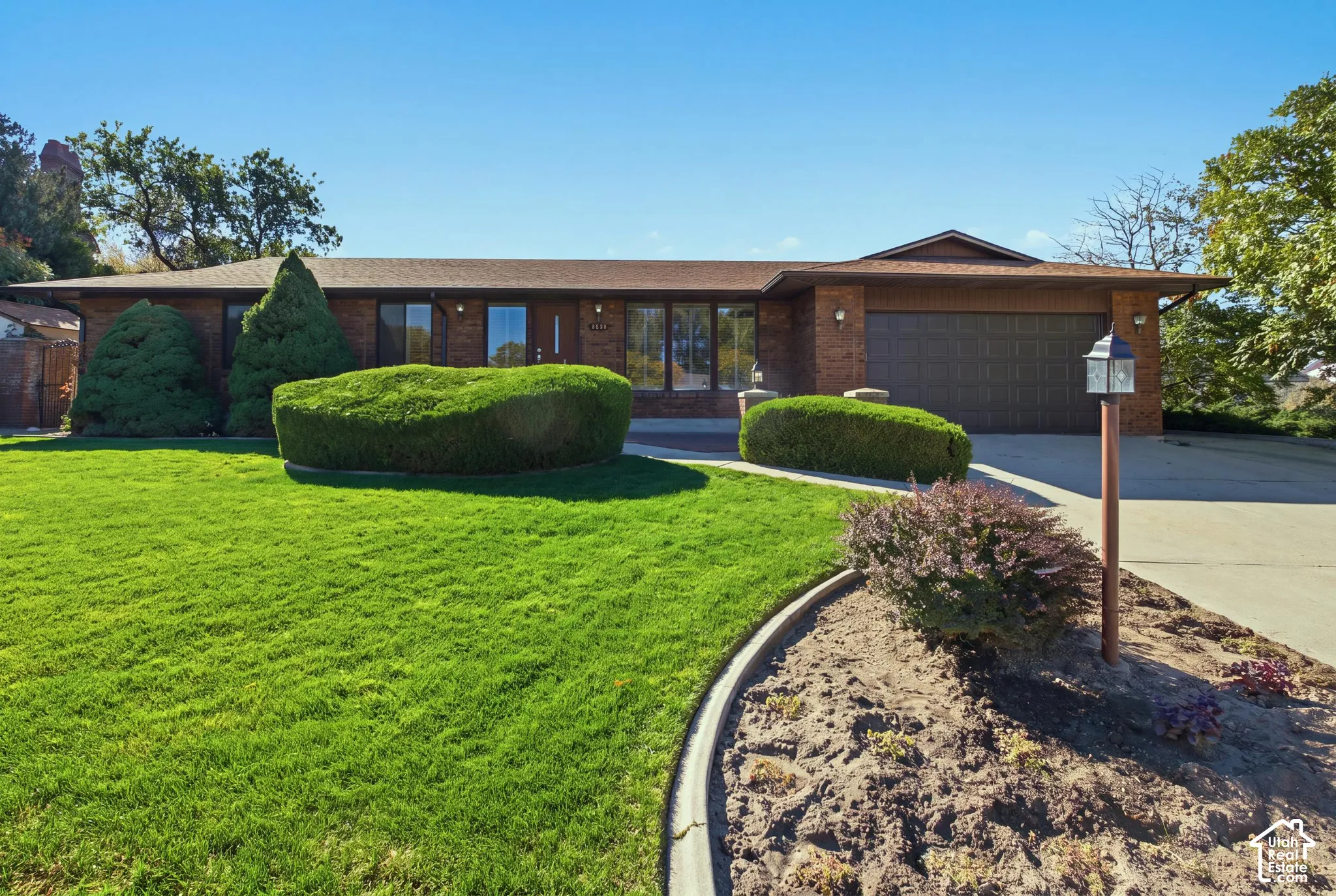 Brick exterior with a brown roofline, attached garage, concrete driveway, and front lawn with mature shrubbery