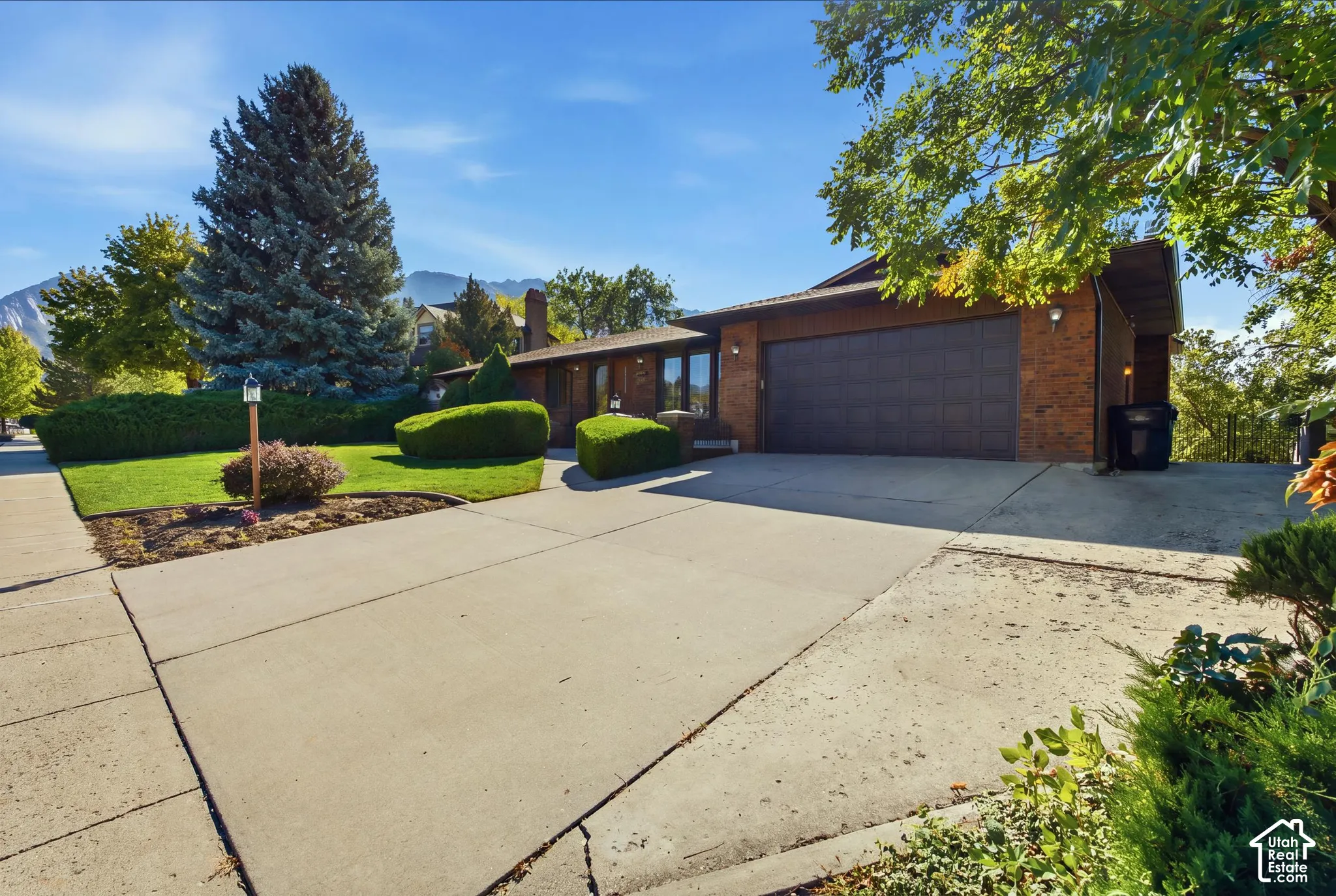Brick exterior featuring a two-car garage, concrete driveway, and landscaped front yard with mature trees