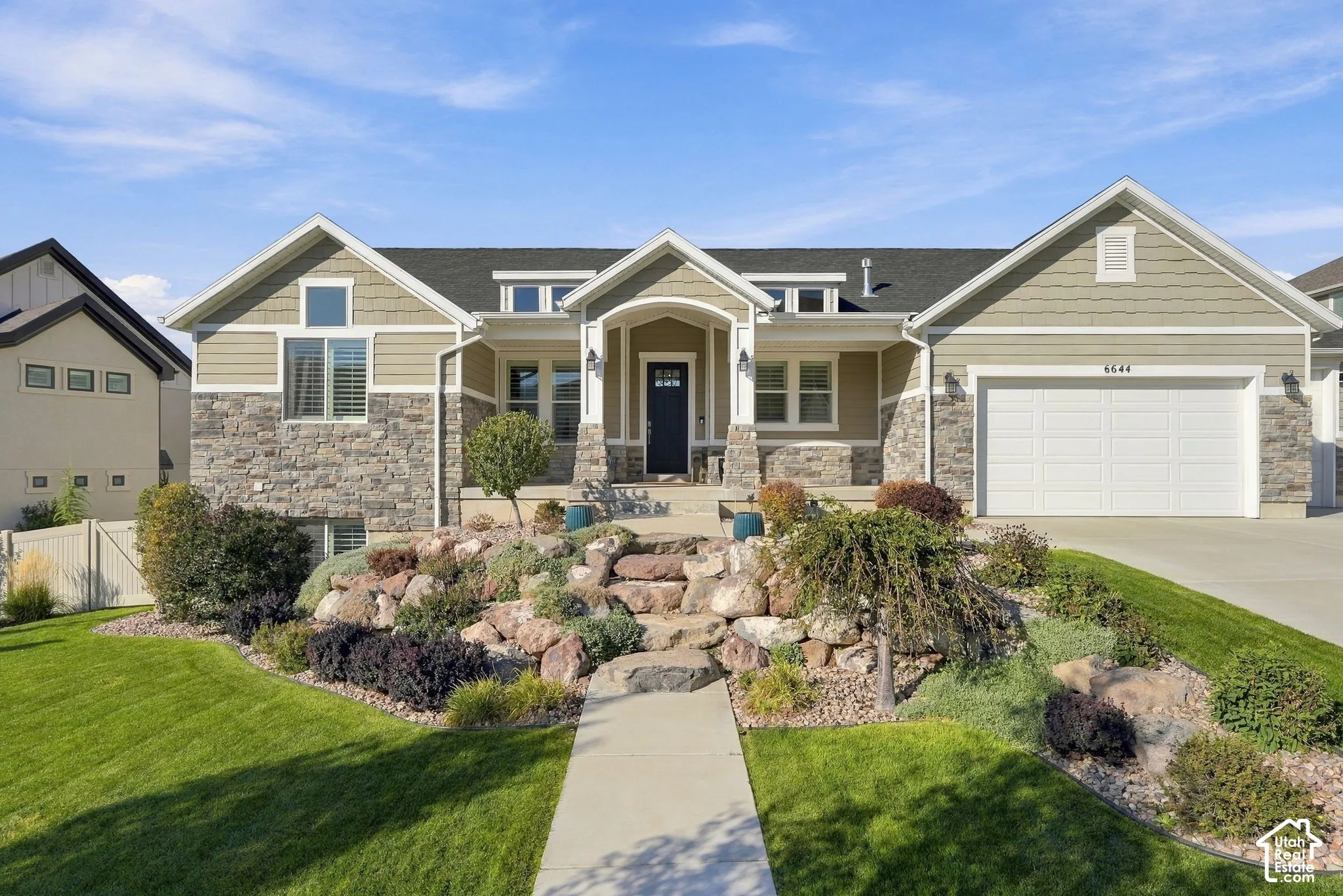 Stone and siding exterior with a multi-level rock garden, mature shrubs, and a concrete pathway leading to the front porch.