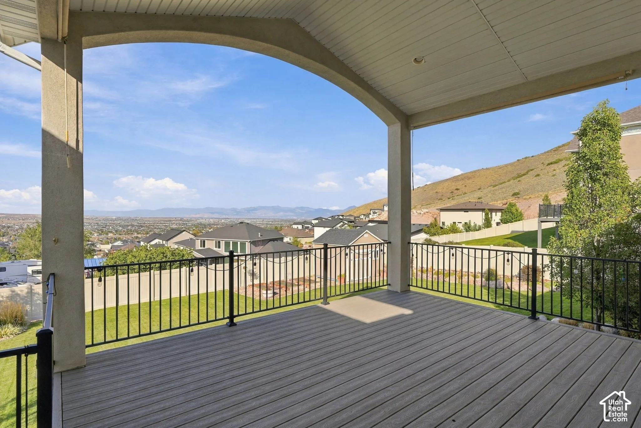 Expansive covered deck featuring composite decking, an arched concrete support, and black metal railings overlooking a landscaped yard and distant terrain