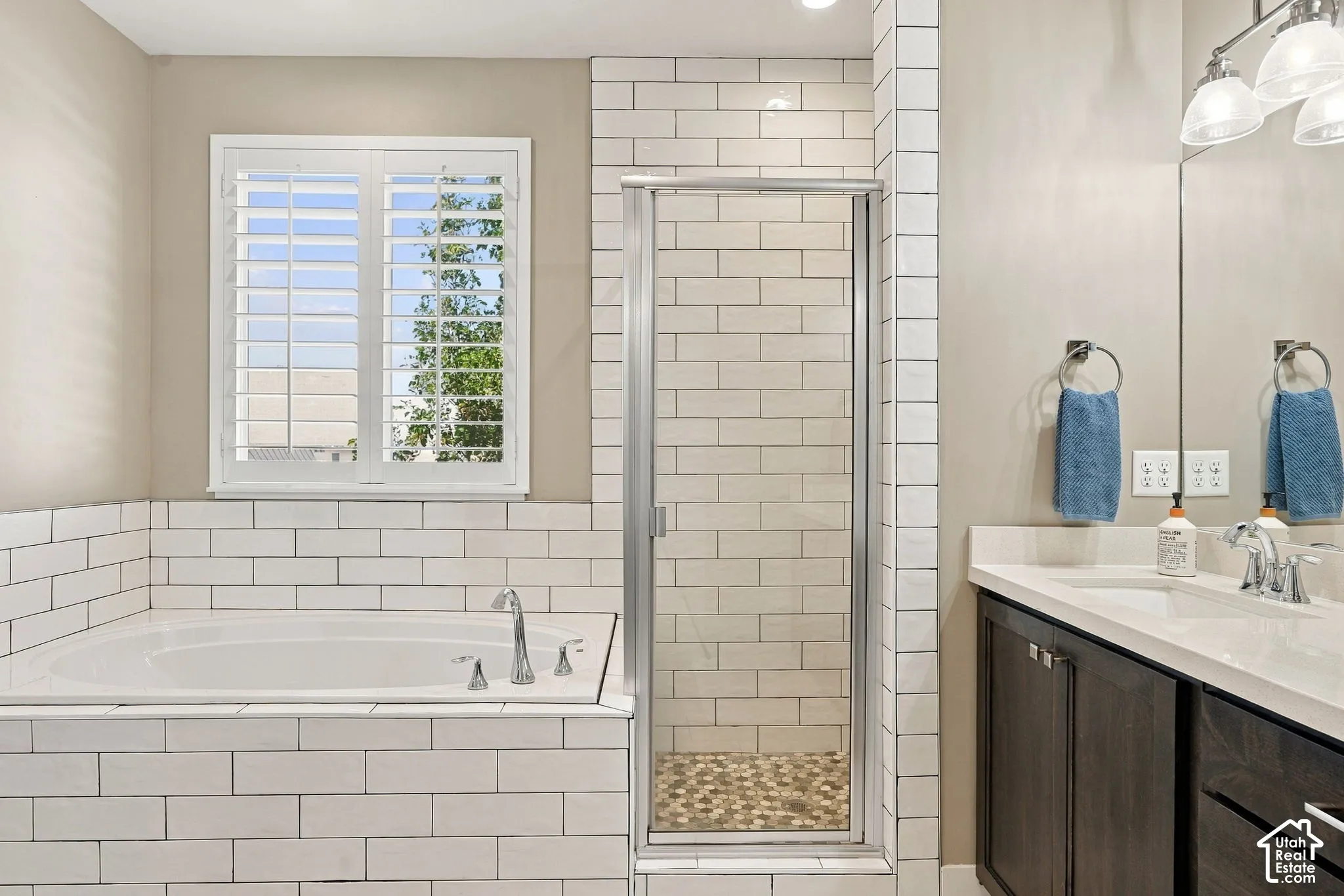Corner soaking tub with subway tile surround, separate glass-enclosed shower with subway tile walls and pebble floor, single vanity with integrated sink, and white window shutters