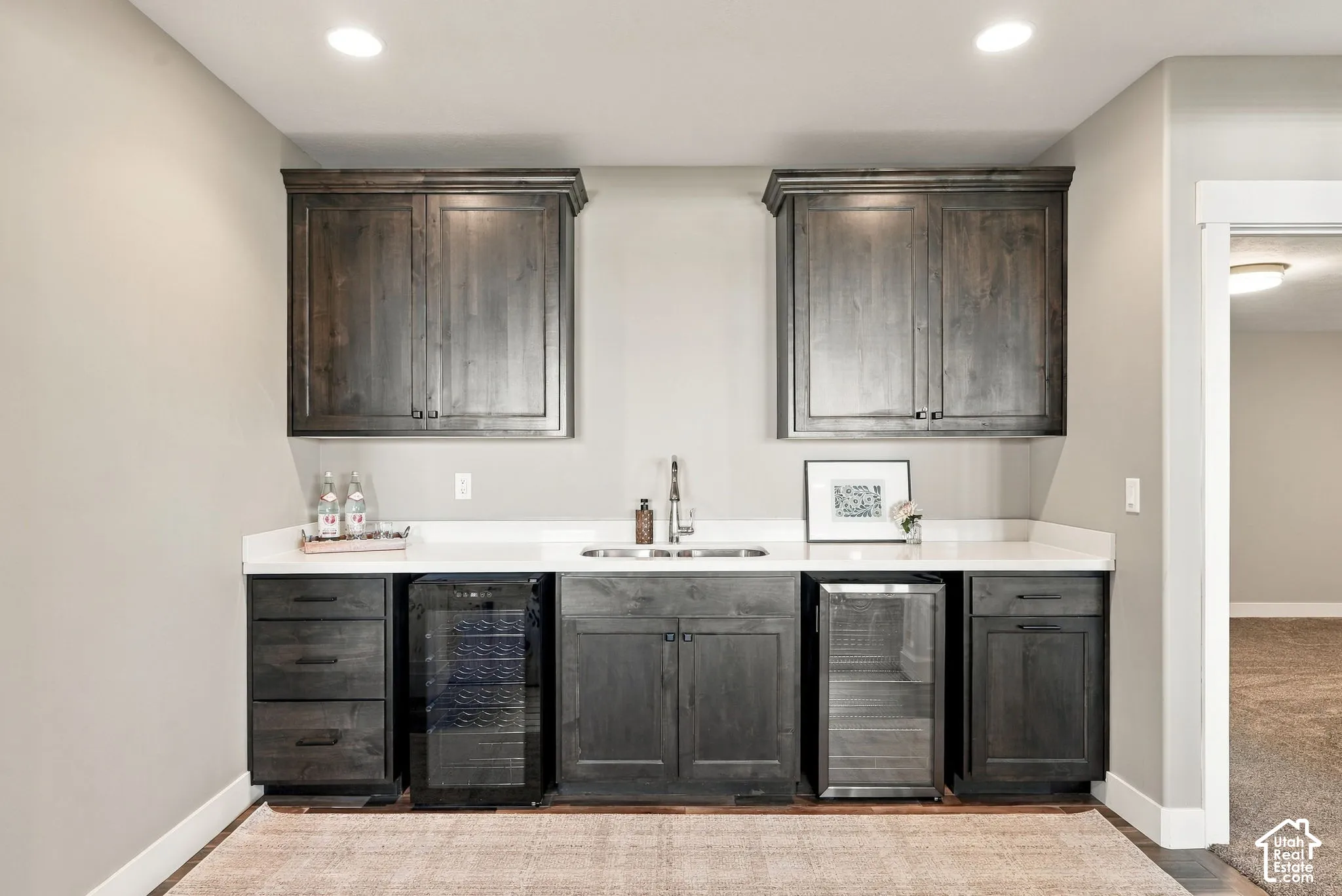 Basement built-in wet bar featuring dark wood-finish cabinetry, white countertops, dual beverage refrigerators, and a stainless steel sink with a gooseneck faucet