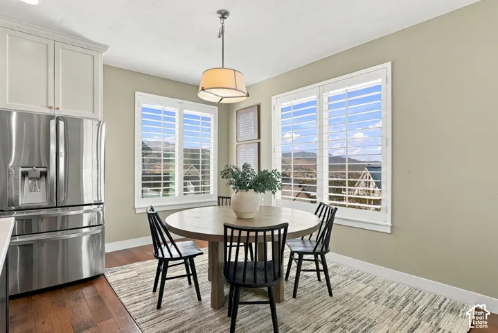 Stainless steel refrigerator, white shaker-style upper cabinetry, wood-finish flooring, two large windows with white louvered shutters, and a drum pendant light fixture
