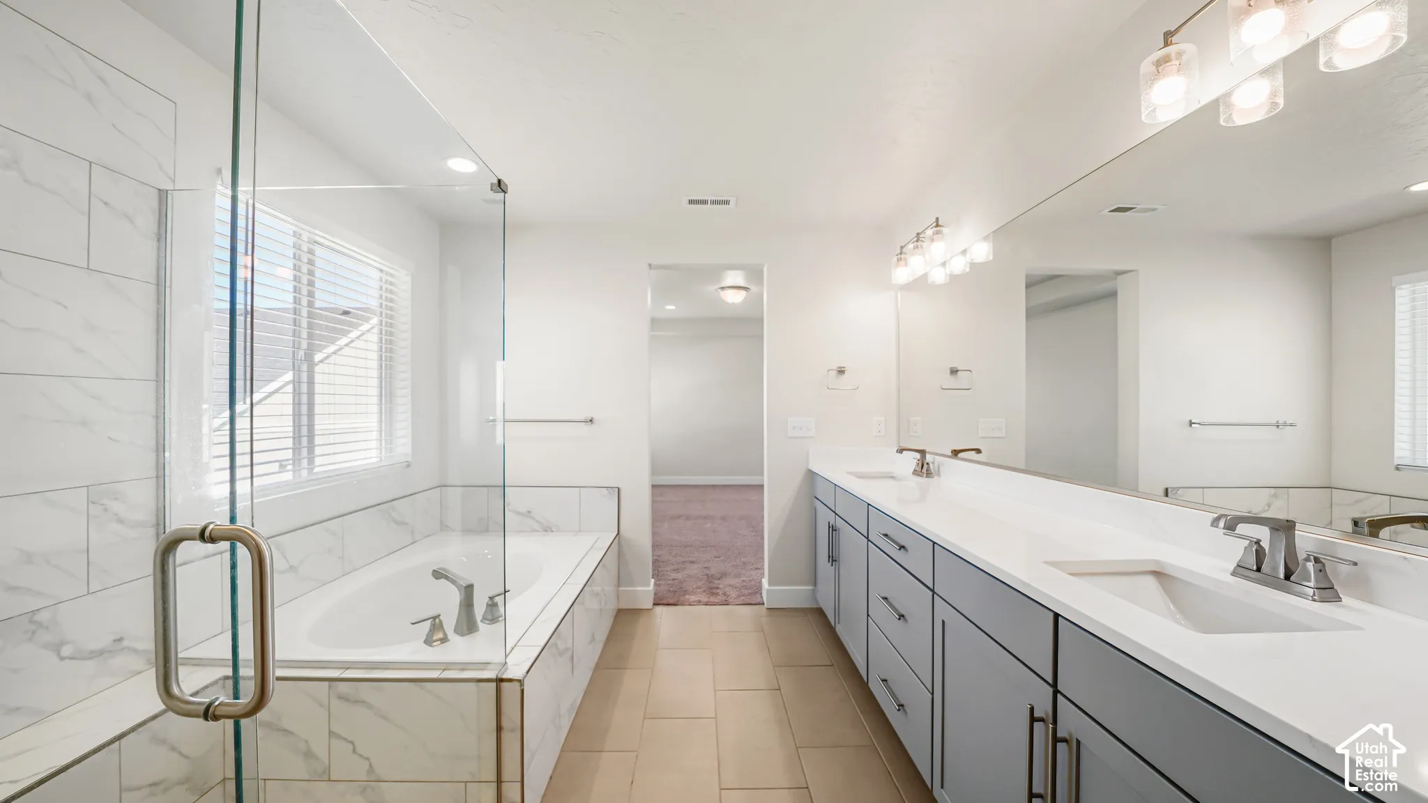 Dual vanity with undermount sinks and light gray cabinetry, large wall-mounted mirror, multiple light fixtures, separate walk-in shower with glass enclosure, and a built-in bathtub with tile surround