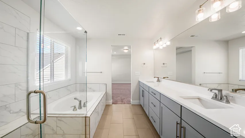Dual vanity with undermount sinks and light gray cabinetry, large wall-mounted mirror, multiple light fixtures, separate walk-in shower with glass enclosure, and a built-in bathtub with tile surround