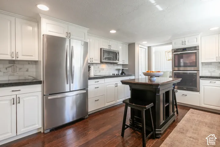 White cabinetry with dark countertops, wood-finish flooring, stainless steel appliances, marble-look tile backsplash, and a central island with built-in storage