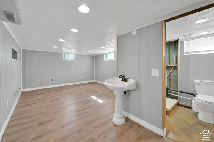 Recessed lighting, wood-finish flooring, light gray wall paneling, white pedestal sink with dark-toned faucet, and a toilet