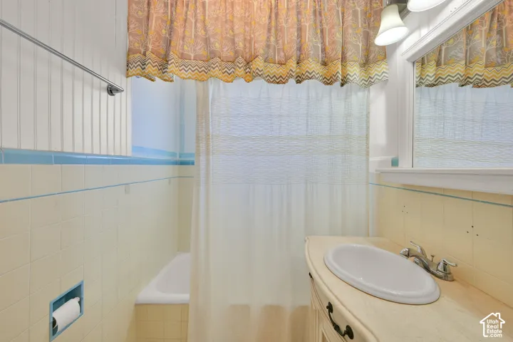 Bright bathroom featuring a pedestal sink with chrome fixtures, ceramic tile walls with blue and cream horizontal striping, a shower-tub combination with clear glass enclosure, recessed ceiling lighting, and decorative valance window treatments providing