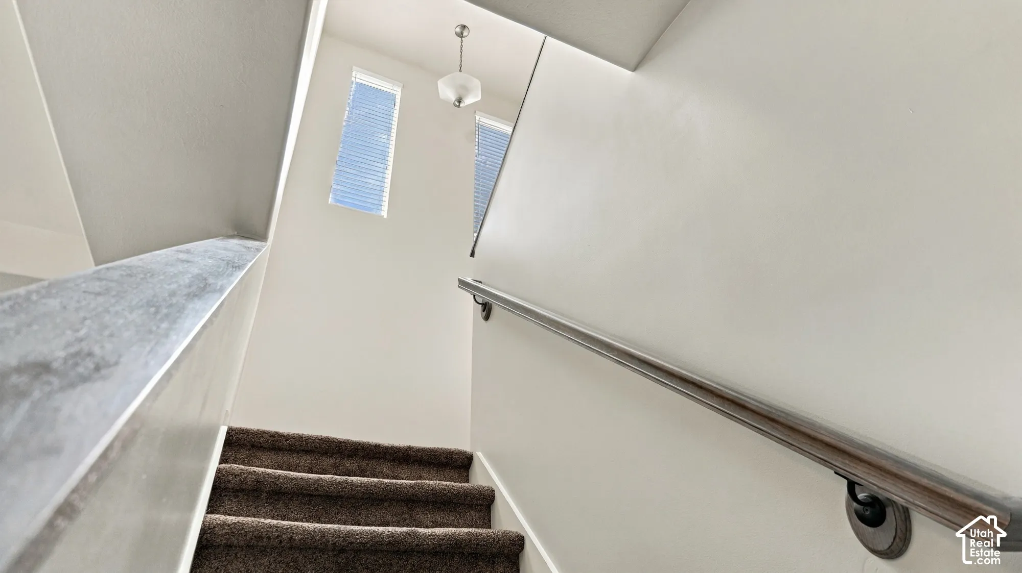 Staircase featuring carpeted treads, a wood-finish handrail, white walls, and upper windows with horizontal blinds