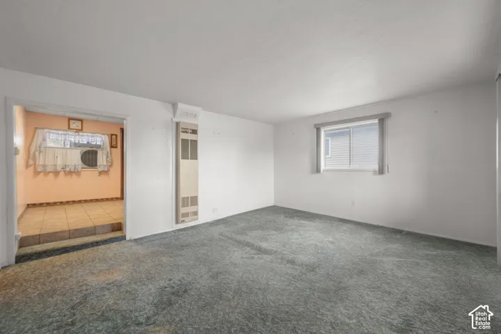 Spacious room with plush carpeting, wall-mounted heating unit, and a window for natural light