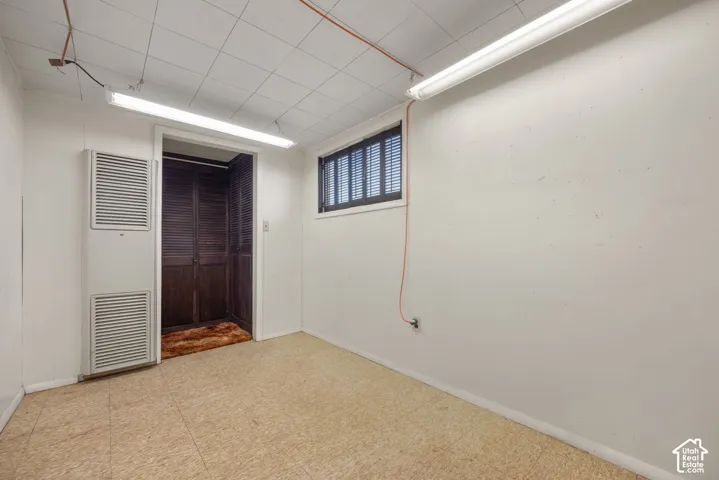 Light-colored flooring with a drop ceiling, white walls, and a window with dark horizontal blinds
