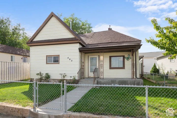 Exterior featuring light-colored siding, a dark brown roof, a covered front porch, and a chain-link fence enclosing a green lawn