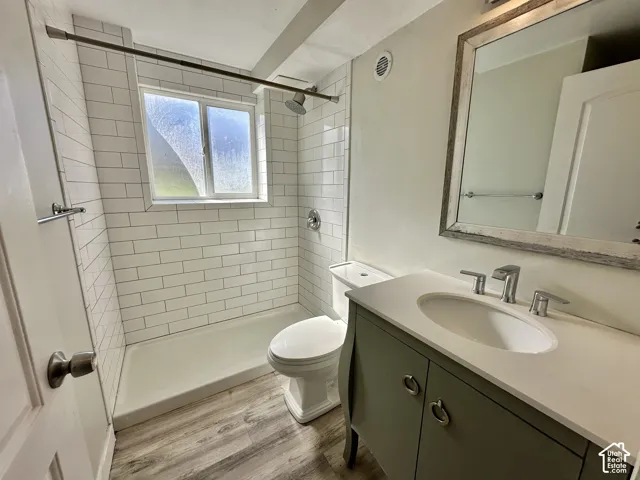 Bathroom featuring wood-finish flooring, a shower with white subway tile surround, a built-in window, a vanity with a white countertop and integrated sink, and a framed mirror