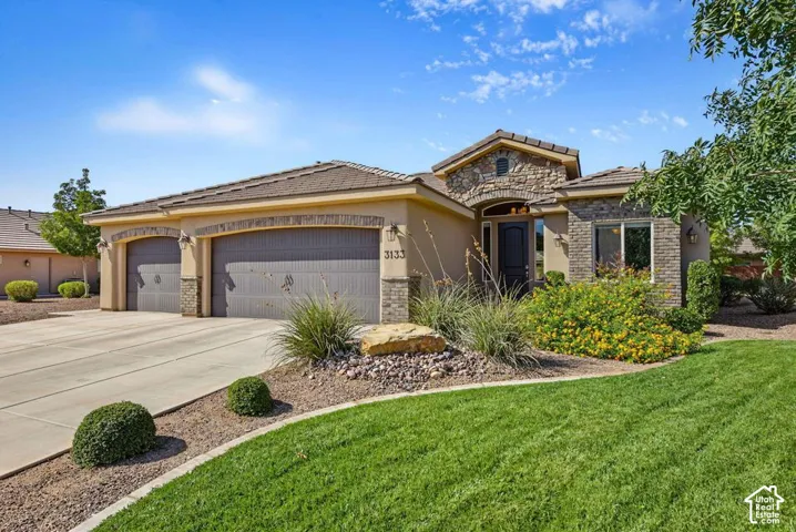 Stucco and stone exterior with a tile roof, featuring a two-car garage, a concrete driveway, and established landscaping including turf and shrubs