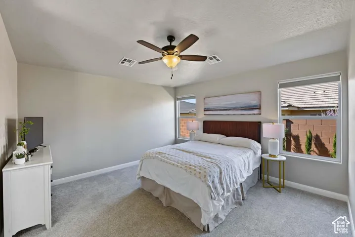 Neutral tone wall paint, textured ceiling, ceiling fan with light fixture, two windows, and light gray carpet flooring