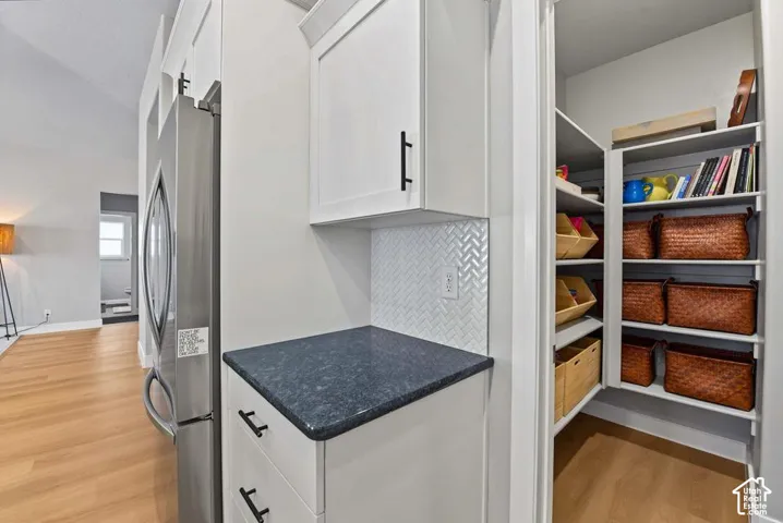 Built-in stainless steel refrigerator, white cabinetry with black hardware, dark countertop, white herringbone tile backsplash, and a walk-in pantry with shelving