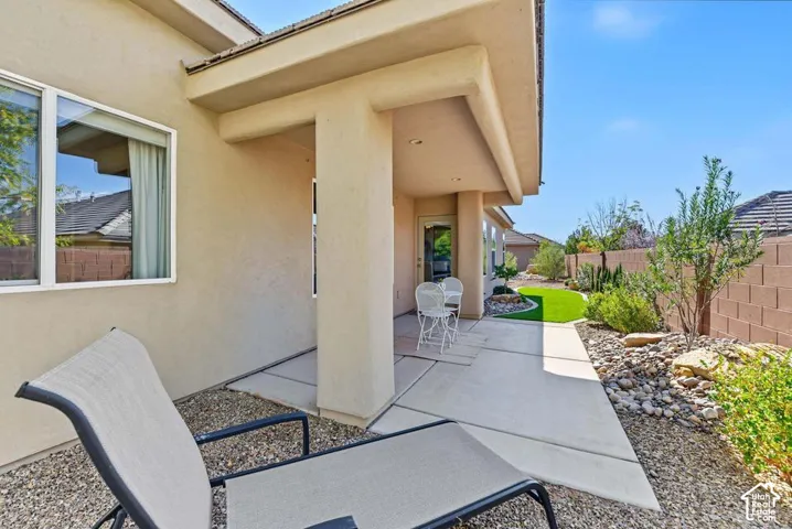 Covered patio with concrete flooring, stucco exterior, and a landscaped yard featuring artificial turf, mature plantings, and a block wall perimeter