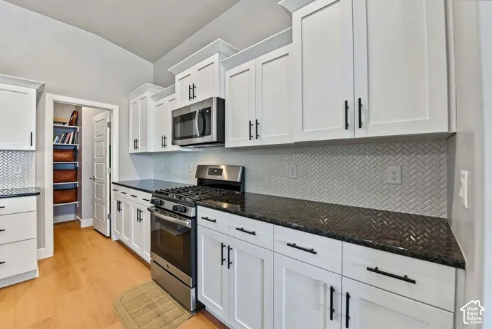 White cabinetry with matte black hardware, stainless steel appliances, dark countertops, a herringbone tile backsplash, and wood-finish flooring