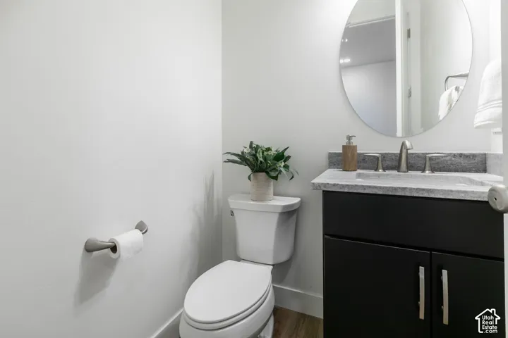 White walls, wood-finish flooring, toilet, dark cabinet vanity with a light countertop, and a circular mirror