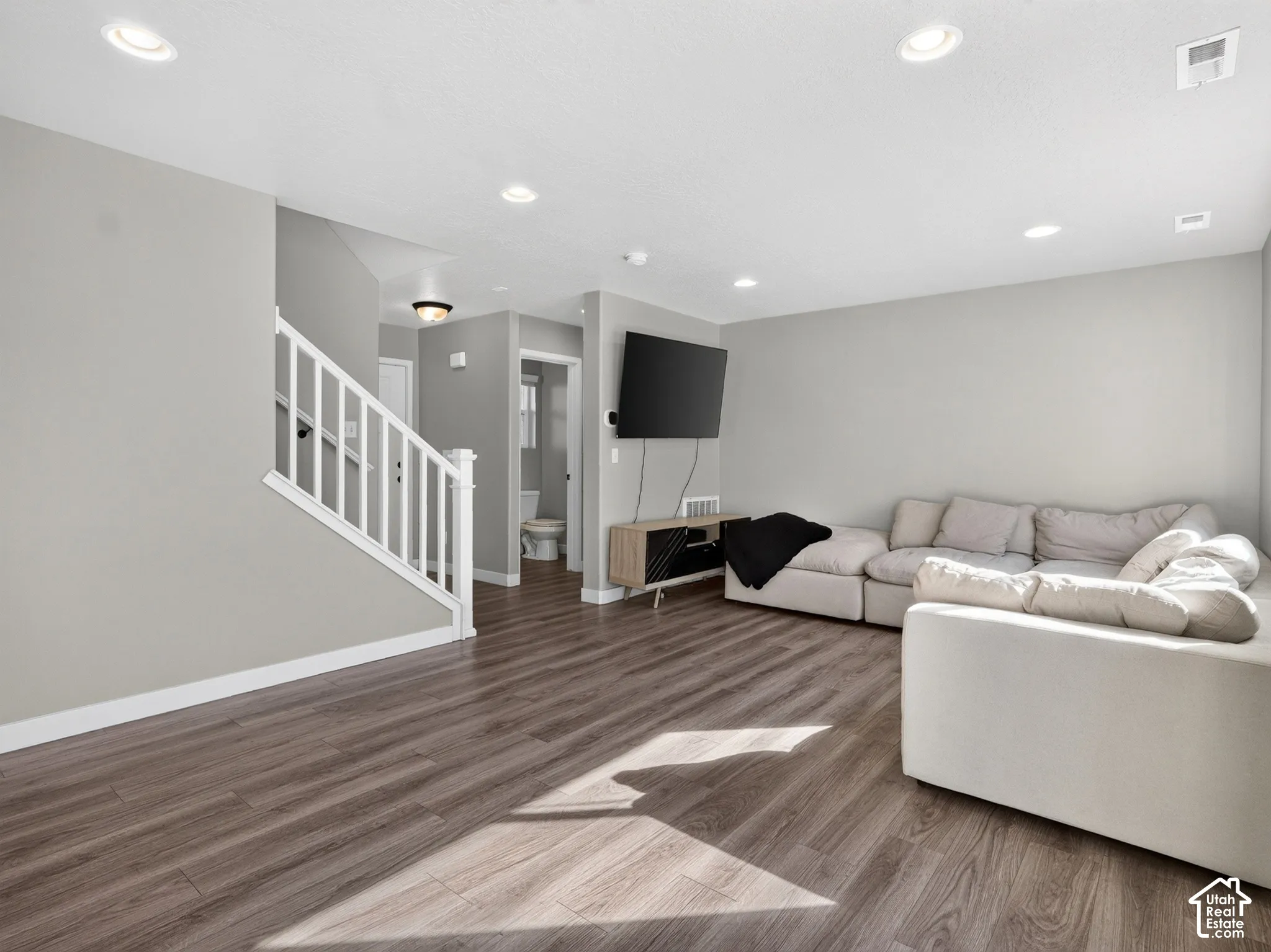 Wood-finish flooring, recessed ceiling lighting, light gray wall finishes, white stair railing, and a white toilet