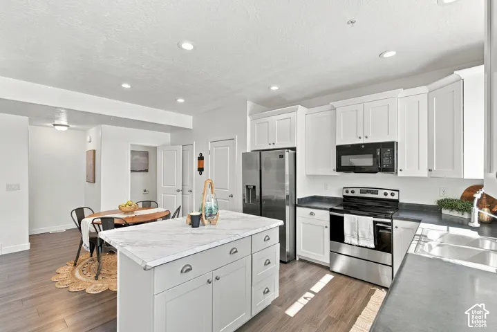 Kitchen featuring white shaker cabinetry, stainless steel appliances, a center island with a light-toned countertop, dark-toned perimeter countertops, and wood-finish flooring
