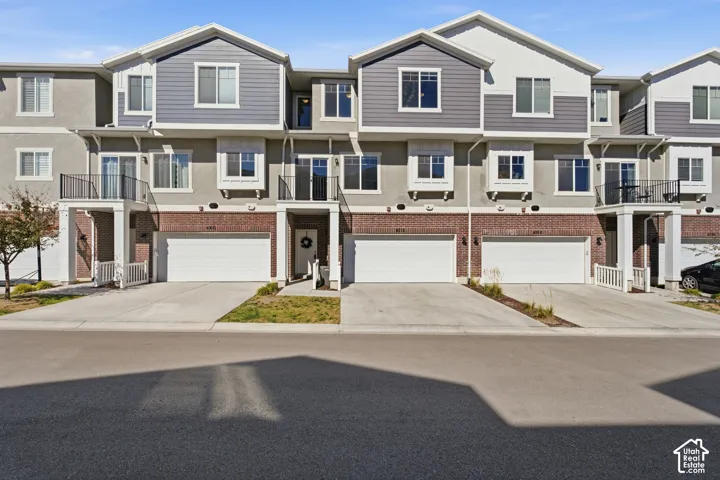 Multi-level townhome exterior featuring an attached garage, brick facade, and a second-story balcony with black railings