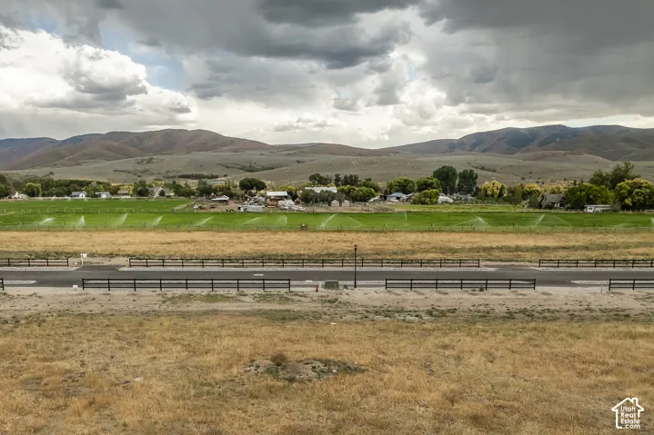 Rural landscape featuring a paved road with black fencing, expansive fields, and distant mountainous terrain
