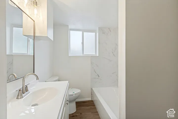 Bathroom featuring a white vanity with an integrated sink, brushed nickel faucet, wood-finish flooring, a white toilet, and a white bathtub with a marble-look tile surround