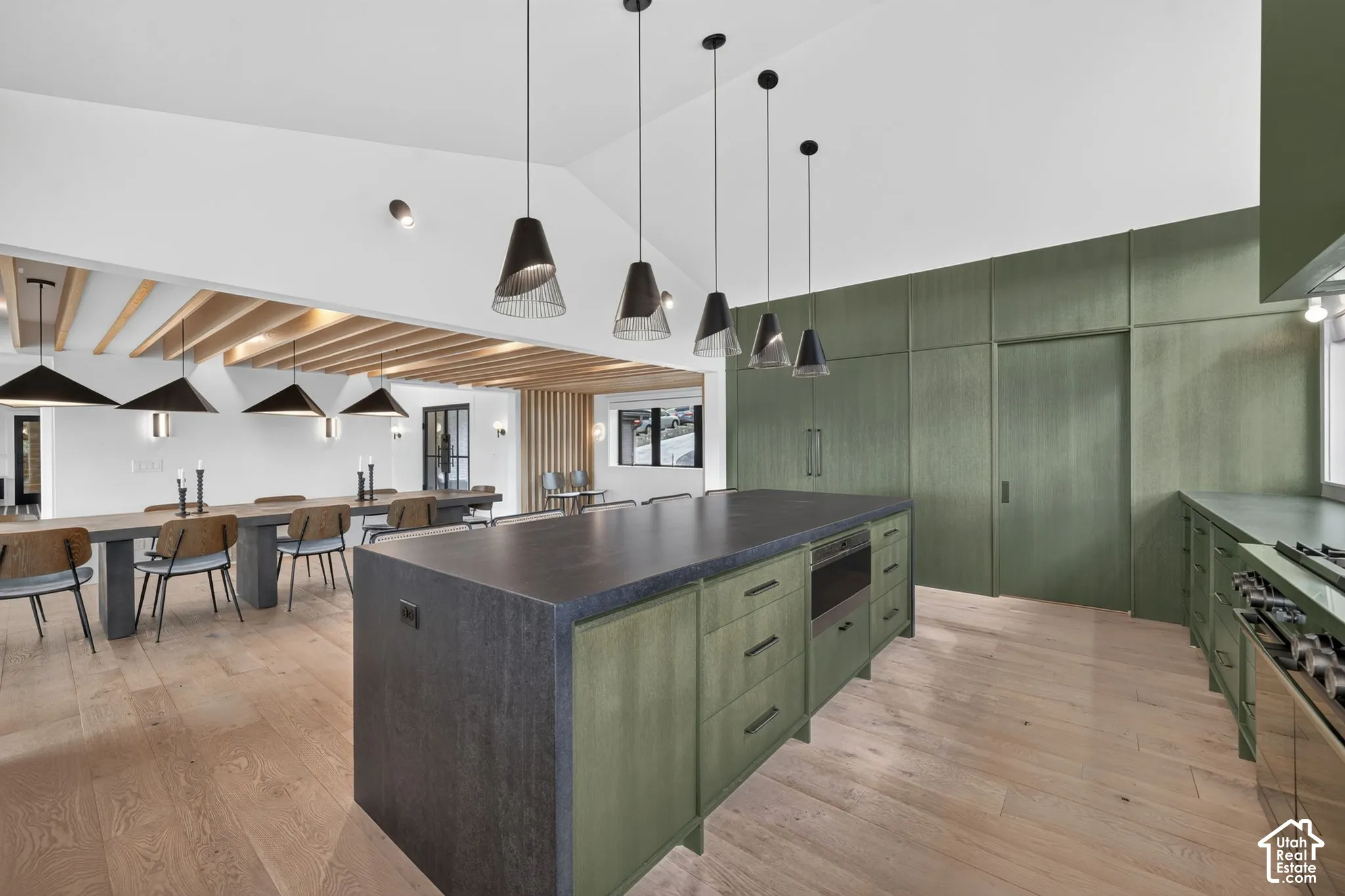 Vaulted ceilings with exposed wood beams, custom green cabinetry, a central island with a built-in oven, light wood-finish flooring, and pendant lighting