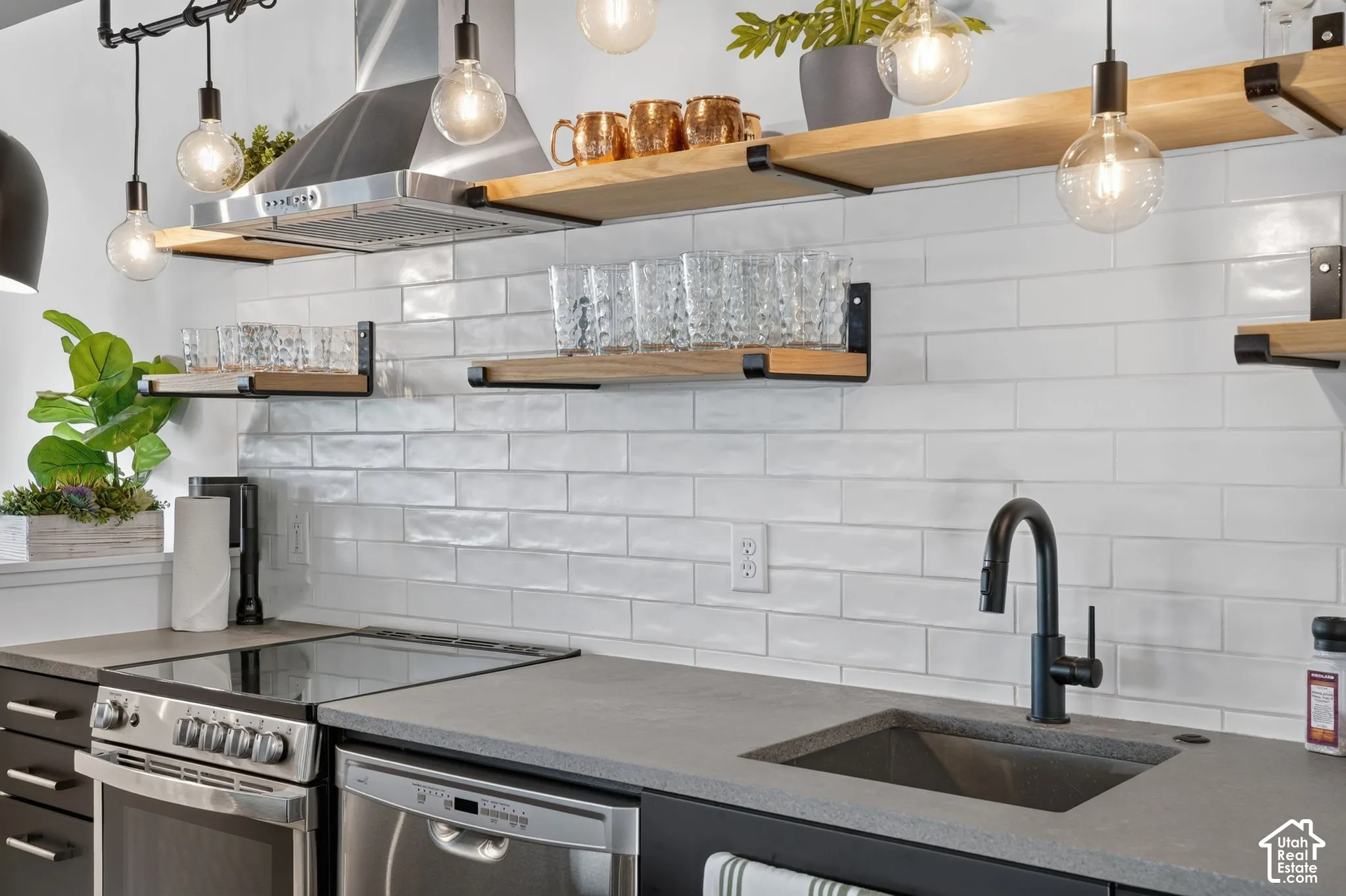 White subway tile backsplash, open wood shelving with black metal supports, stainless steel range hood, stainless steel dishwasher, and dark composite sink with a matte black faucet