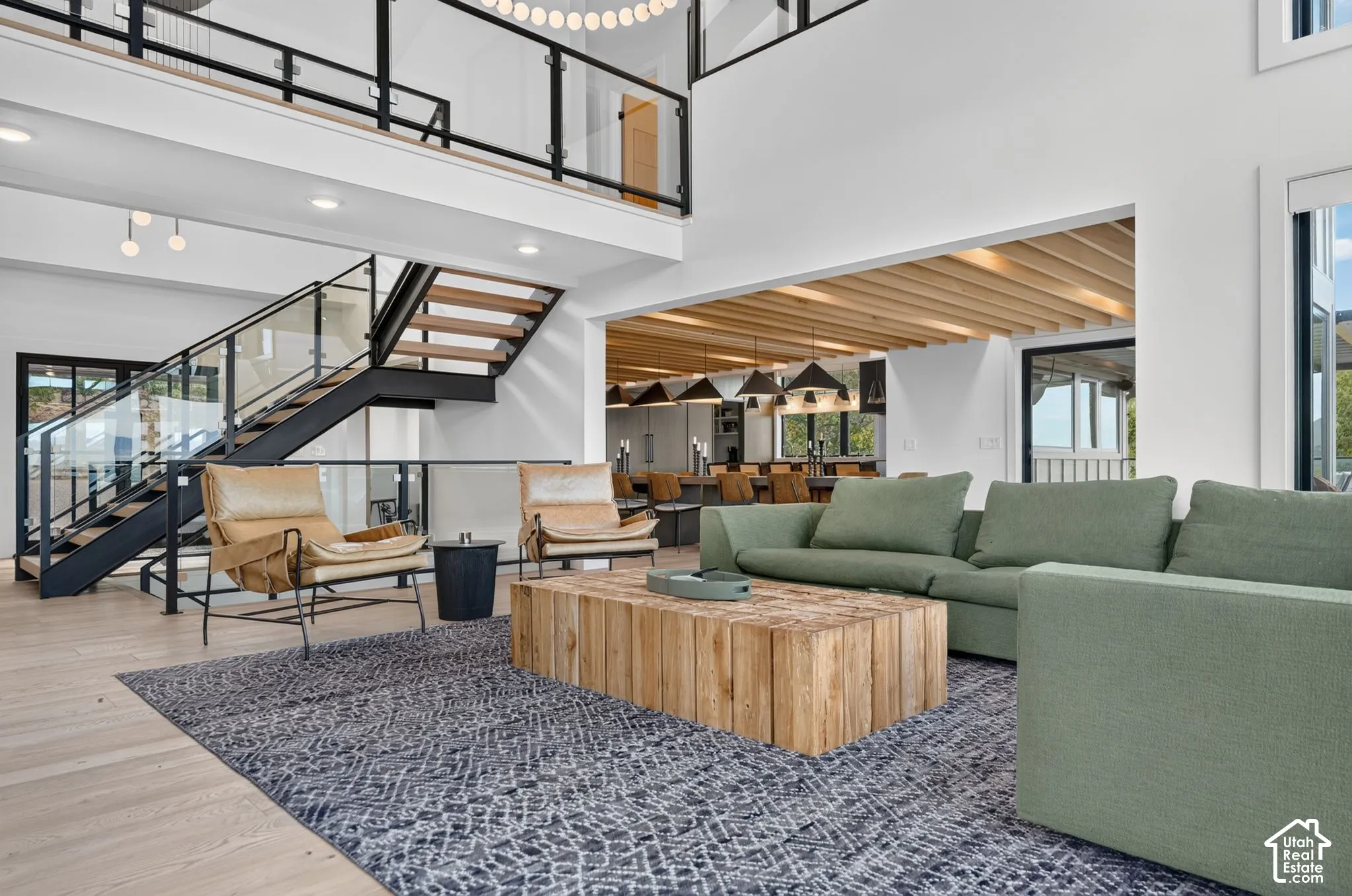 Spacious living area featuring a two-story ceiling, a modern open-tread staircase with glass and metal railings, wood-finish flooring, and exposed wood beam detailing