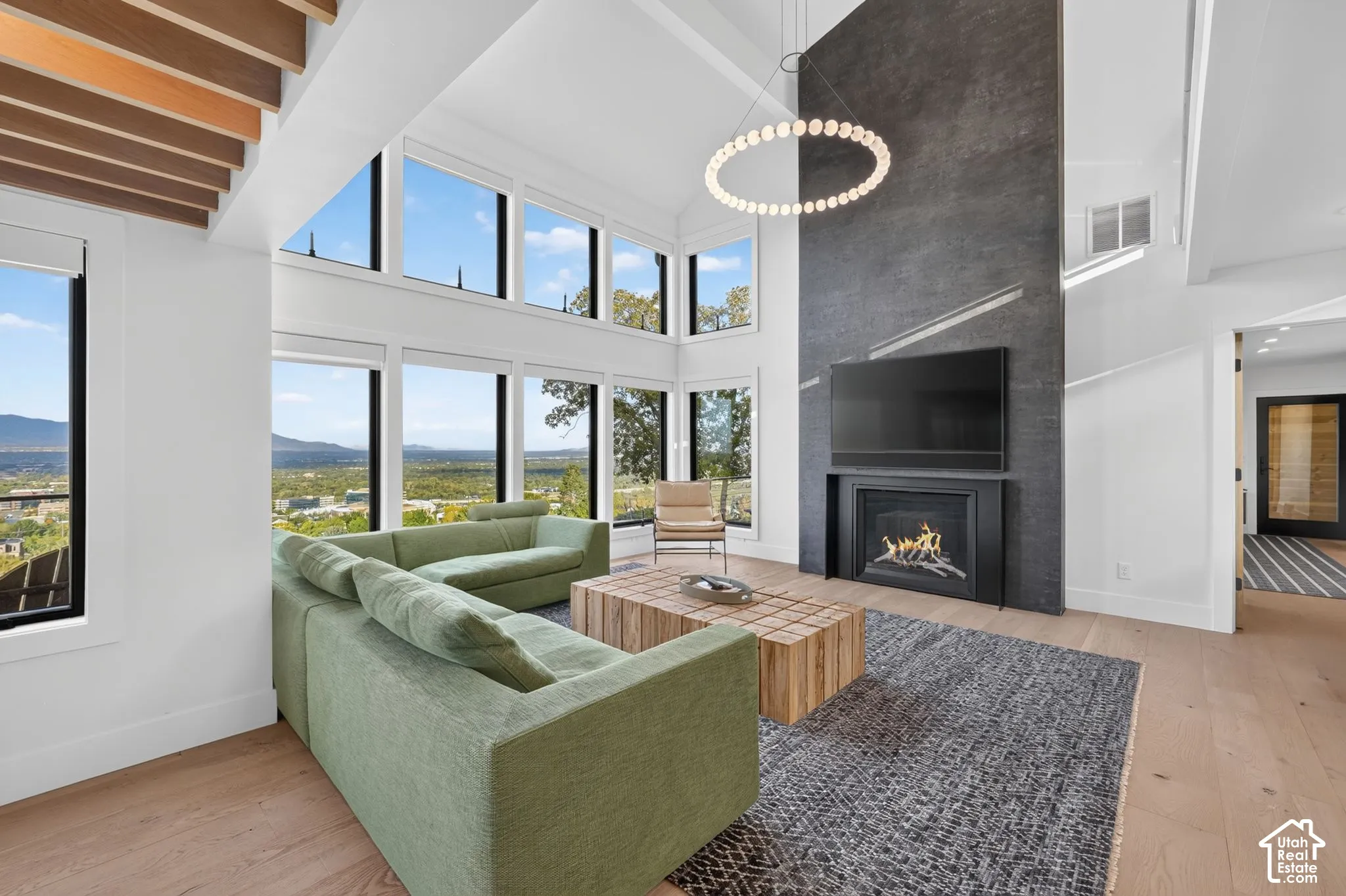 Vaulted ceilings with exposed wood beams, expansive floor-to-ceiling windows, modern fireplace with a dark stone surround, wood-finish flooring, and a contemporary circular chandelier